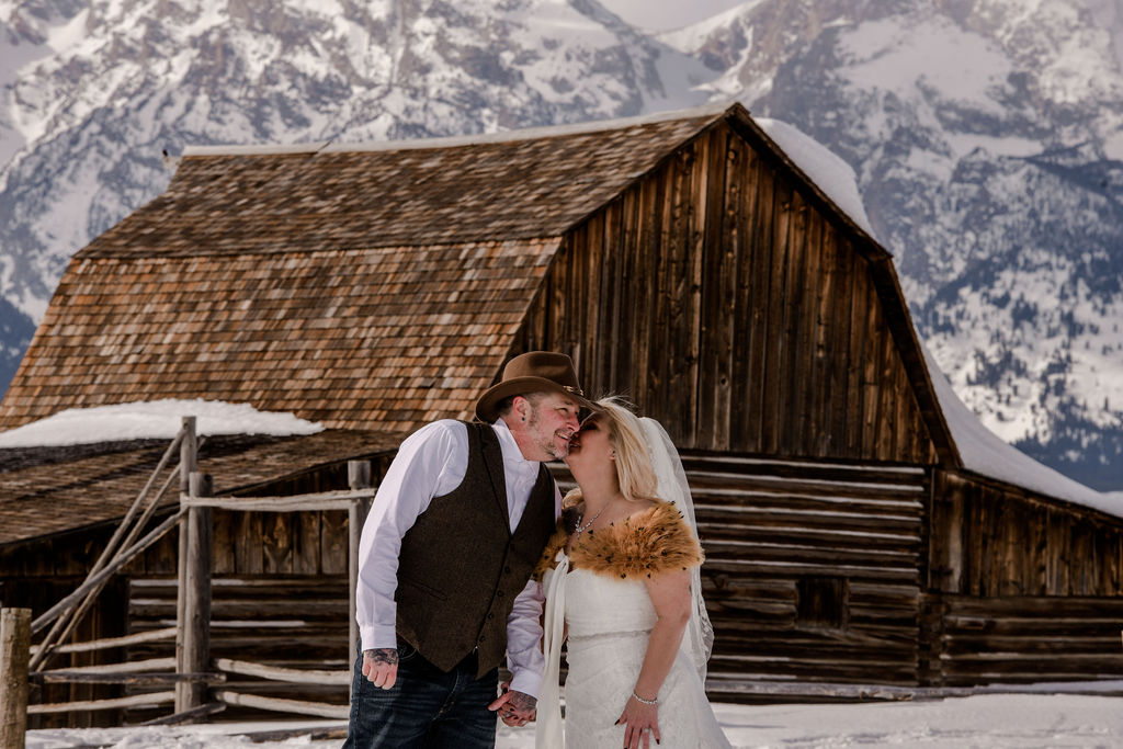 couple kiss in front of snowy Moulton Barns