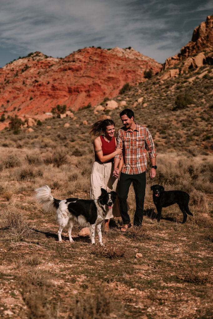 couple walk in a field with their dogs with the red rock in the background